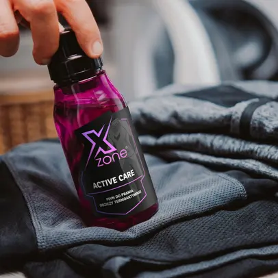 Butelka Xzone Active Care – płyn do prania odzieży termoaktywnej, trzymana w dłoni obok sportowych ubrań.